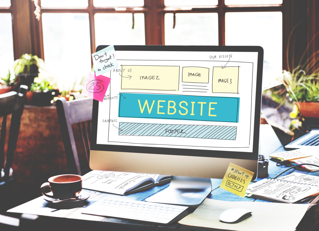 Monitor shows a website wireframe layout on a cluttered desk with sticky notes; plants and a notebook in the background.