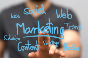Person writing blue handwritten marketing terms on a glass surface: Social, Web, Marketing, Trends, Content, Strategy, Media (blurred background)